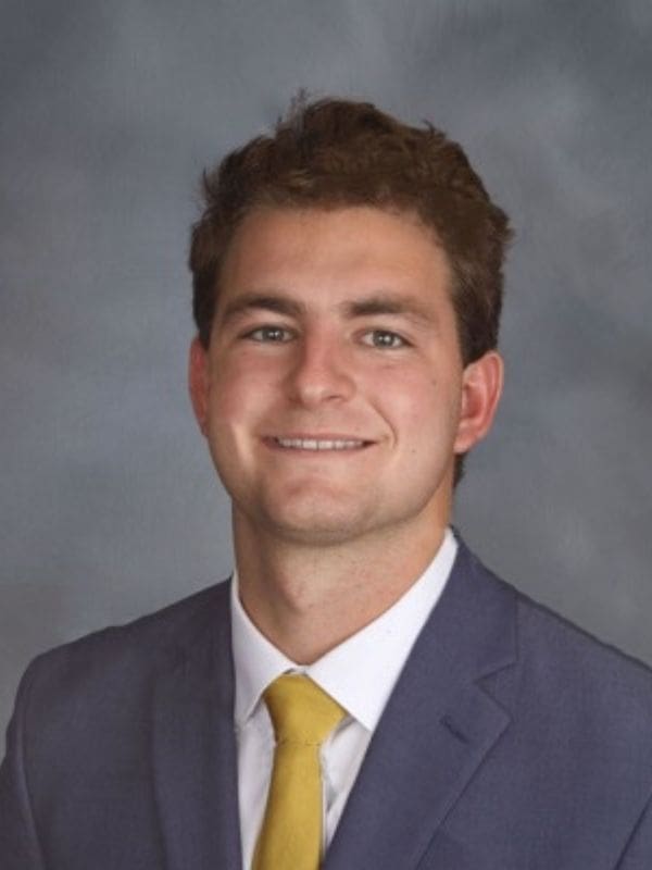 A photo of Tyler Totin wearing a white dress shirt, a golden tie, and a dark gray suit.
